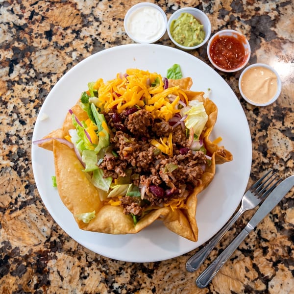 Crispy tortilla bowl topped with seasoned ground beef, beans, lettuce, tomatoes, onions, and shredded cheddar, served with dipping sauces.