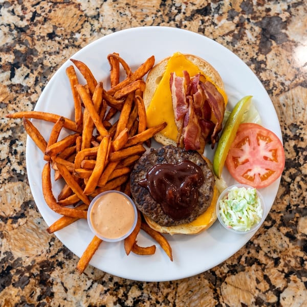 Plate of barbecue cheeseburger with bacon, sweet potato fries, tomato, pickle, and coleslaw on a granite countertop.